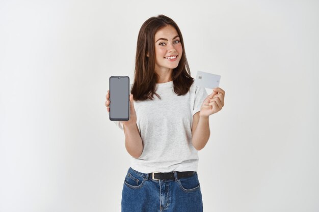 Portrait d'une jeune fille heureuse aux longs cheveux bruns debout sur un mur blanc, montrant une carte de crédit en plastique et un écran de smartphone vierge.