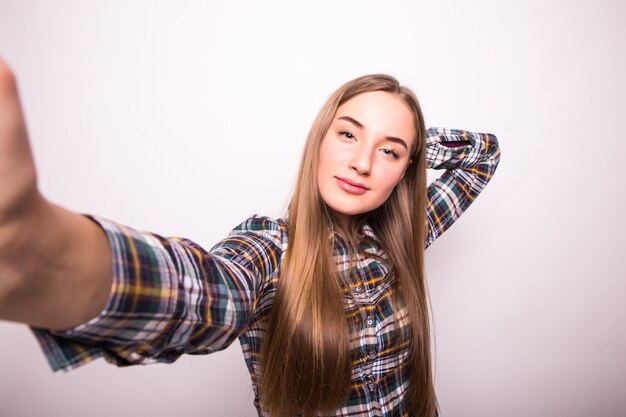 Portrait d'une jeune fille heureuse faisant la grimace tout en prenant des photos d'elle-même isolé sur mur blanc