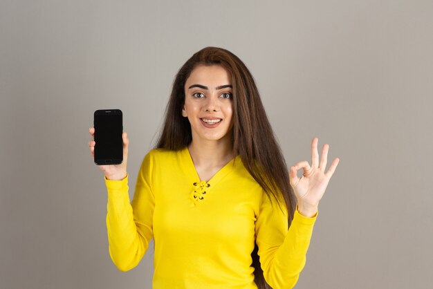 Portrait de jeune fille en jaune tenant un téléphone portable sur un mur gris.