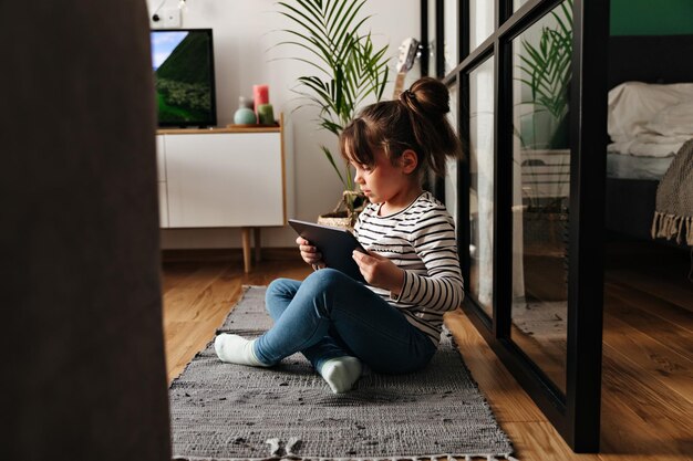 Portrait de jeune fille en jeans assis sur un tapis dans la chambre et jouant sur une tablette