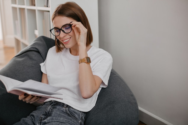 Portrait de jeune fille à lunettes et T-shirt blanc avec sourire livre de lecture dans une chaise grise.