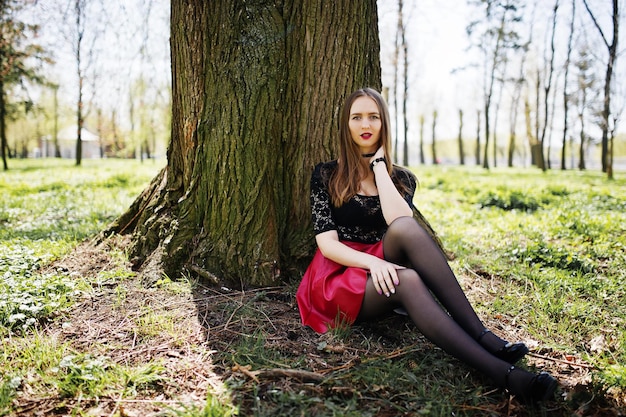 Portrait de jeune fille avec un maquillage lumineux avec des lèvres rouges collier tour de cou noir sur son cou et une jupe en cuir rouge assis près d'un arbre au parc