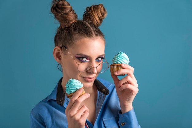 Portrait de jeune fille à la mode dans des verres ronds avec maquillage professionnel et coiffure chignon sur fond bleu. la fille tient deux cupcakes. fermer. copiez l'espace.