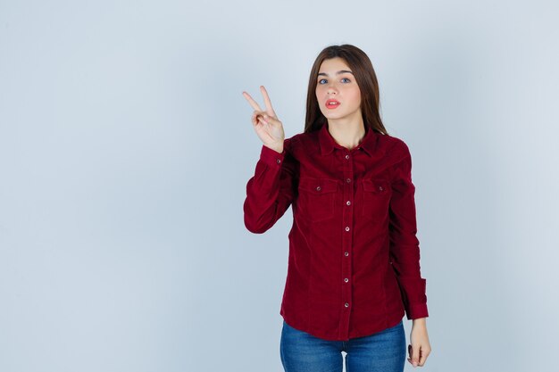 Portrait de jeune fille montrant un geste de victoire en blouse bordeaux et à la perplexité