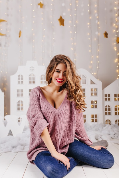 Portrait de jeune fille en pull violet avec col en V assis sur le sol dans un mur lumineux avec des guirlandes et des maisons de jouets blanches.