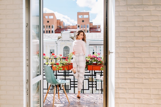 Portrait jeune fille en pyjama se déplaçant sur balcon en ville en matinée ensoleillée. Ses longs cheveux volant au vent, elle sourit.