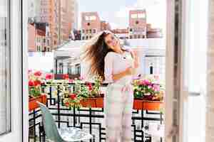 Photo gratuite portrait jeune fille en pyjama se déplaçant sur balcon en ville en matinée ensoleillée. ses longs cheveux volant au vent, elle sourit.