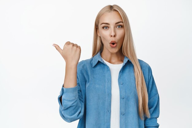 Portrait d'une jeune fille à la recherche de produits curieux pointant du doigt vers la gauche et regardant fixement intrigué en posant une question debout sur fond blanc