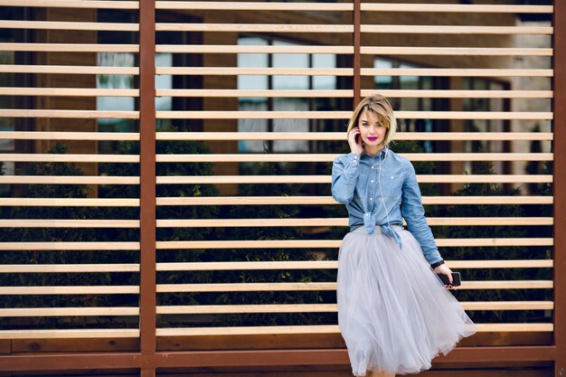 Portrait d'une jeune fille rêveuse avec des cheveux blonds courts et des lèvres rose vif, écouter de la musique sur un smartphone avec des arcs en bois rayés derrière