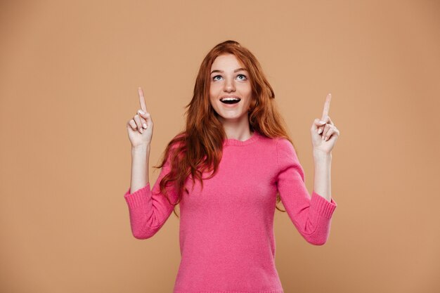 Portrait d'une jeune fille rousse joyeuse pointant vers le haut avec les doigts