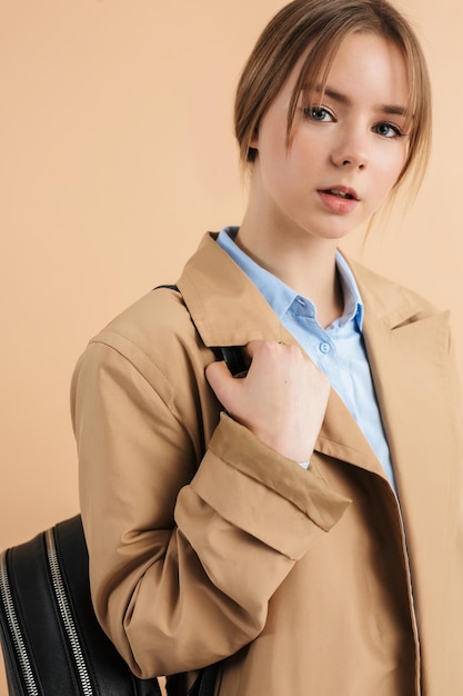Portrait de jeune fille séduisante en trench-coat et chemise bleue avec sac à dos sur l'épaule tout en regardant rêveusement à huis clos sur fond beige