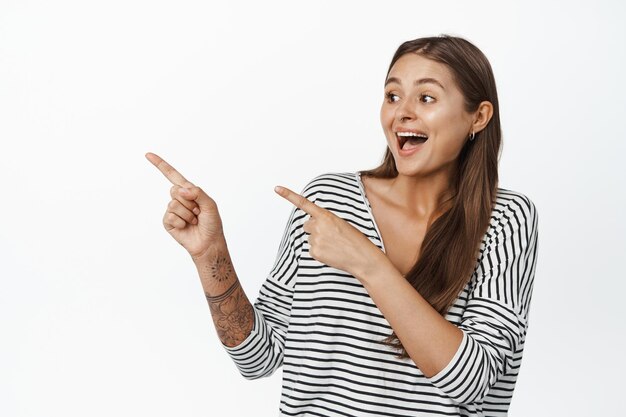 Portrait d'une jeune fille souriante excitée vérifiant une grande vente, pointant les doigts vers la gauche et riant étonné, debout sur fond blanc.