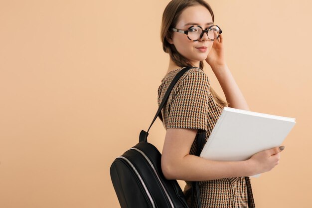 Portrait de jeune fille souriante séduisante avec deux tresses en combinaison de tweed et lunettes avec sac à dos noir sur l'épaule tenant un livre à la main tout en regardant joyeusement à huis clos sur fond beige