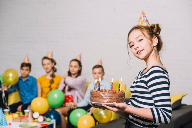 Portrait d&#39;une jeune fille souriante tenant un gâteau d&#39;anniversaire avec des amis à l&#39;arrière-plan