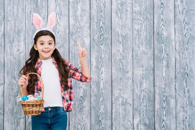 Portrait d&#39;une jeune fille tenant le panier d&#39;oeufs de Pâques pointant le doigt vers le haut sur un fond en bois