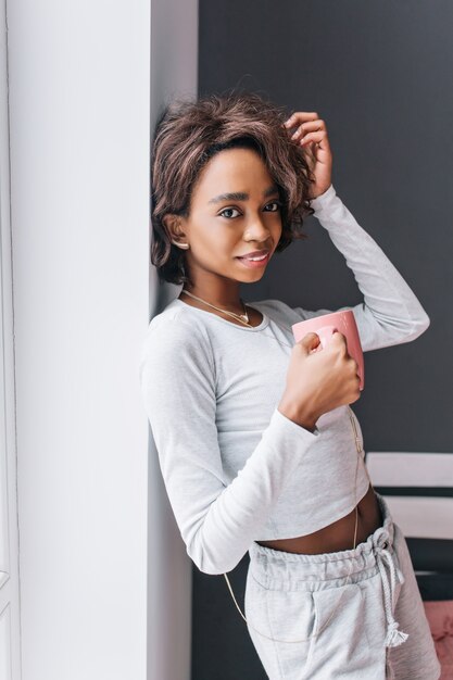 Portrait de jeune fille, tenant une tasse rose, appréciant le café du matin, boire du thé, souriant dans la chambre avec un mur gris sur l'espace