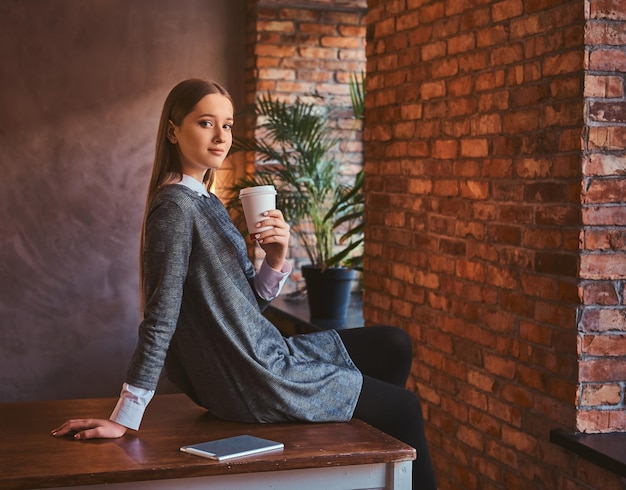 Portrait d'une jeune fille vêtue d'une élégante robe grise tient une tasse de café à emporter en regardant une caméra tout en étant assise sur une table dans une pièce avec un intérieur loft.