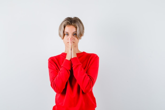 Portrait d'un jeune garçon adolescent avec les mains en geste de prière en pull rouge et à la vue de face pleine d'espoir