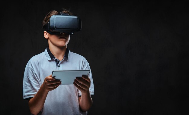 Portrait d'un jeune garçon étudiant vêtu d'un t-shirt blanc à l'aide de lunettes de réalité virtuelle et d'une tablette. Isolé sur un fond sombre.