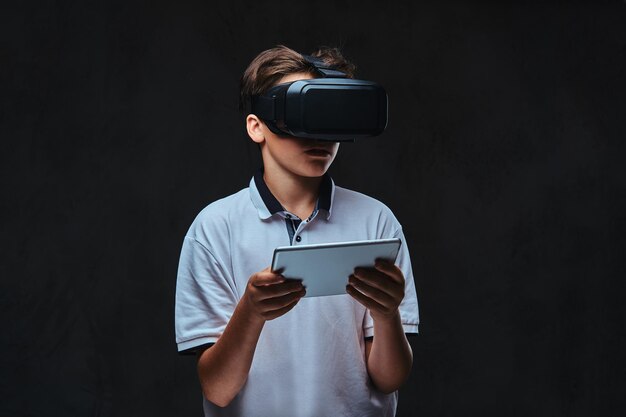 Portrait d'un jeune garçon étudiant vêtu d'un t-shirt blanc à l'aide de lunettes de réalité virtuelle et d'une tablette. Isolé sur un fond sombre.