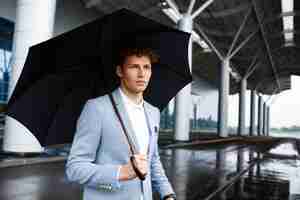 Photo gratuite portrait de jeune homme d'affaires aux cheveux roux tenant un parapluie noir dans la rue des pluies