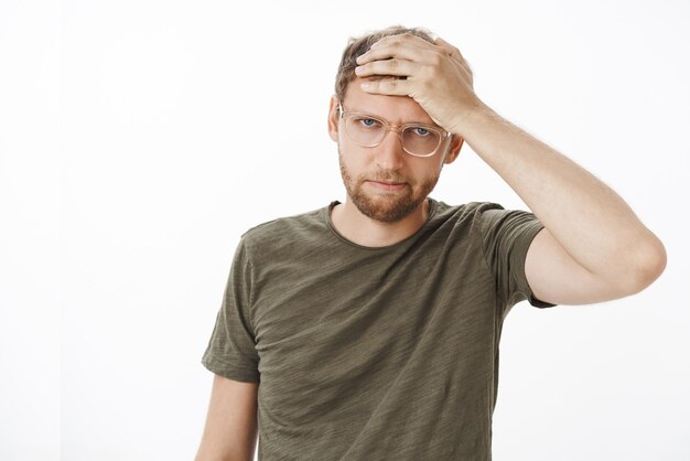 Portrait de jeune homme d'affaires ennuyé et irrité dans des verres et t-shirt vert foncé tenant la main sur le front regardant énervé et en colère perdre son humeur