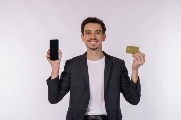 Portrait d'un jeune homme d'affaires heureux debout montrant un téléphone portable et détenant une carte bancaire de crédit isolé sur fond de couleur blanche studio