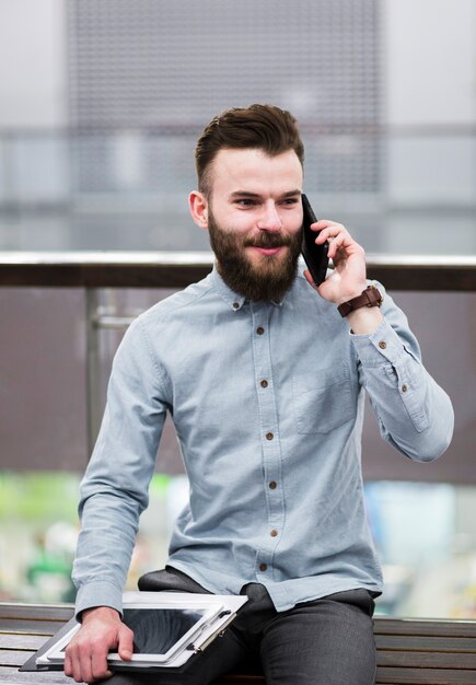 Portrait, jeune, homme affaires, séance banc, tenue, presse-papiers, et, tablette numérique, parler, sur, téléphone portable