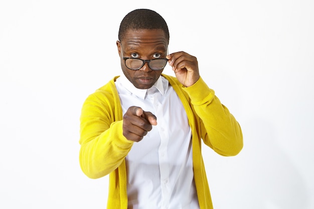 Portrait de jeune homme africain à la mode employé ou étudiant portant une chemise blanche et un gilet jaune regardant par-dessus ses lunettes, pointant la caméra avec une expression faciale difficile, vous choisissant