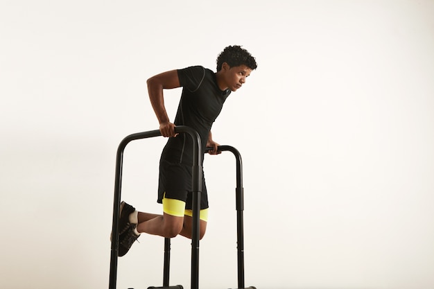 Un portrait d'un jeune homme afro-américain musclé concentré dans des vêtements d'entraînement noirs faisant des trempettes sur des barres parallèles sur blanc