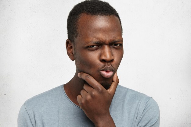 Portrait de jeune homme afro-américain pensif suspect en t-shirt décontracté touchant le visage tout en réfléchissant à quelque chose, en essayant de trouver une solution, ayant une expression perplexe et perplexe