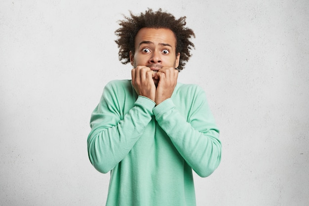 Photo gratuite portrait de jeune homme afro-américain avec peur et expression anxieuse, se mord les ongles, porte un pull vert,