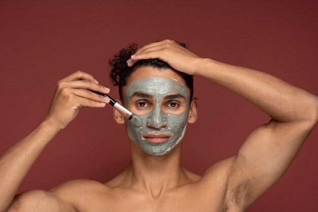Portrait d'un jeune homme appliquant un masque facial sur son visage avec un pinceau de maquillage