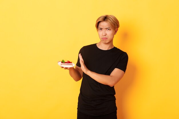 Portrait de jeune homme asiatique dégoûté et bouleversé refuse de manger un gâteau, l'air mécontent et montrant un geste de rejet, mur jaune.