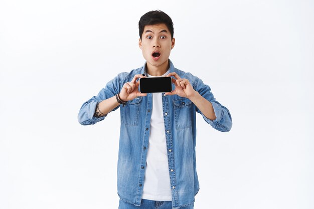 Portrait d'un jeune homme asiatique impressionné et étonné montrant une nouvelle bande-annonce de film sur l'écran d'un smartphone, tenant un téléphone portable horizontalement, la bouche ouverte amusée