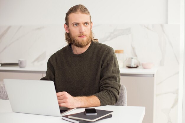 Portrait de jeune homme assis à la table avec un ordinateur portable tout en regardant rêveusement la fenêtre