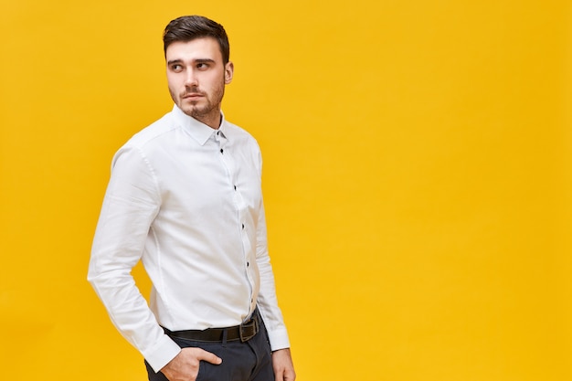 Portrait de jeune homme aux cheveux noirs à la mode avec chaume ayant un regard sérieux, tournant la tête contre le mur jaune blanc avec espace de copie pour votre texte ou informations promotionnelles