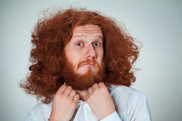 Portrait de jeune homme aux longs cheveux roux et avec une expression faciale choquée sur fond gris