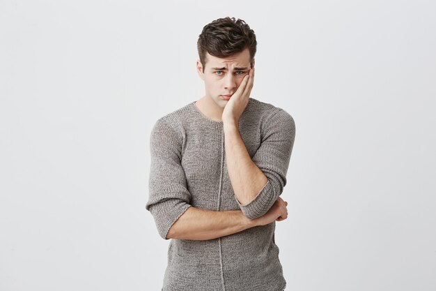 Portrait d'un jeune homme aux yeux bleus triste et bouleversé aux cheveux noirs, portant un pull, gardant la main sur la joue, fronçant les sourcils, regardant avec une expression triste, à cause des mauvaises nouvelles qu'il a reçues.