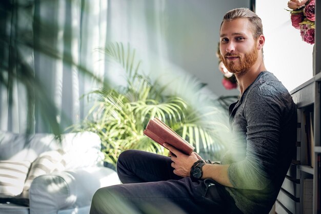 Portrait de jeune homme barbu rousse tenant un livre dans une pièce avec des plantes vertes.