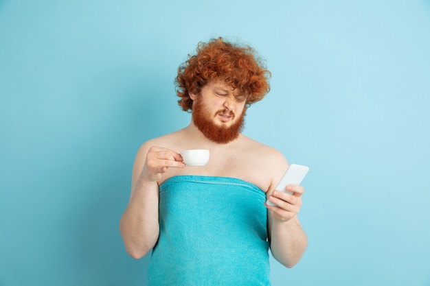 Portrait de jeune homme caucasien dans sa journée de beauté et routine de soins de la peau. Modèle masculin aux cheveux roux naturels, boire du café et regarder les médias sociaux. Soins du corps et du visage, concept de beauté naturelle et masculine.