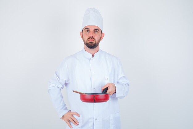 Portrait de jeune homme chef tenant une poêle avec une cuillère en bois en uniforme blanc et à la grave vue de face