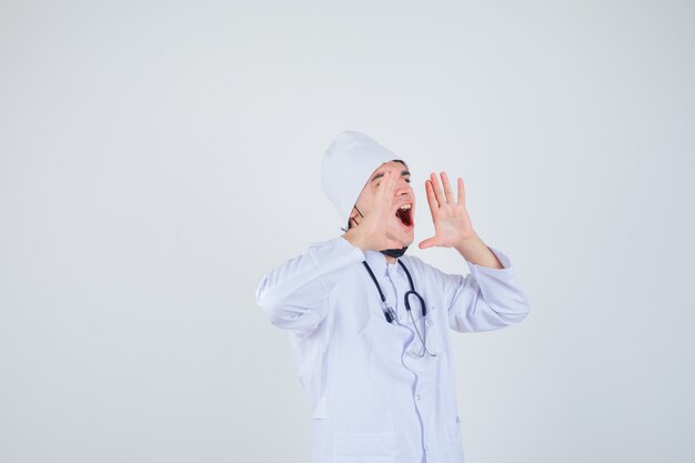 Portrait de jeune homme criant tout en gardant les mains près de la bouche en uniforme blanc, masque et à la colère