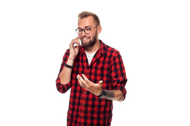 Portrait d'un jeune homme décontracté parlant au téléphone et souriant tout en regardant ailleurs, quelque part en haut. Tourné en studio. Jeune homme hipster en chemise à carreaux et lunettes portant sur fond blanc