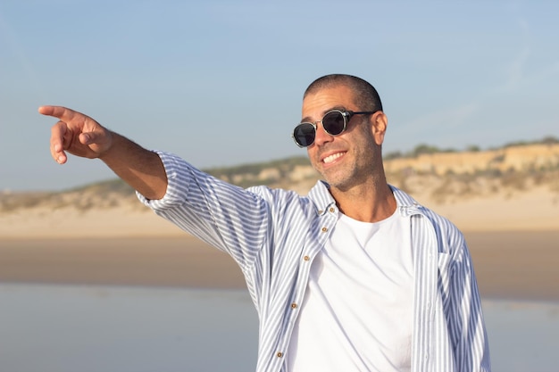 Portrait de jeune homme élégant sur la plage. Modèle masculin avec la tête rasée dans des lunettes de soleil souriant vivement, pointant. Portrait, vacances, concept de beauté