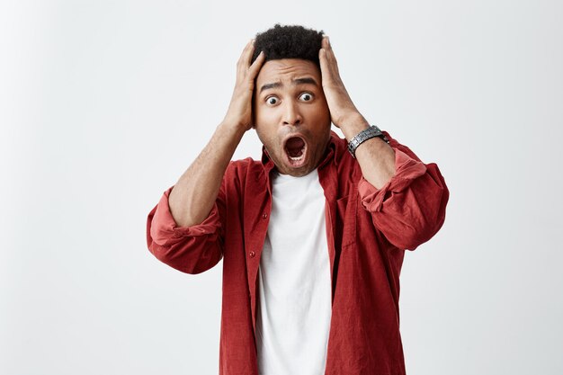 Portrait de jeune homme étonné à la peau sombre avec une coiffure afro en t-shirt blanc sous une chemise rouge tenant la tête avec les mains, regardant à huis clos avec la bouche ouverte et une expression choquée.