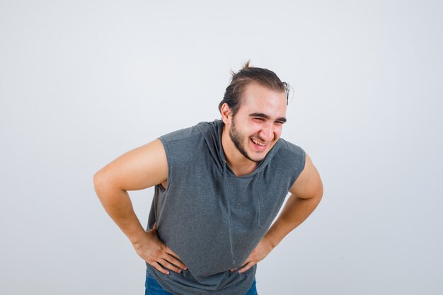 Portrait de jeune homme en forme posant avec les mains sur la taille tout en se penchant en sweat à capuche sans manches et à la joyeuse vue de face