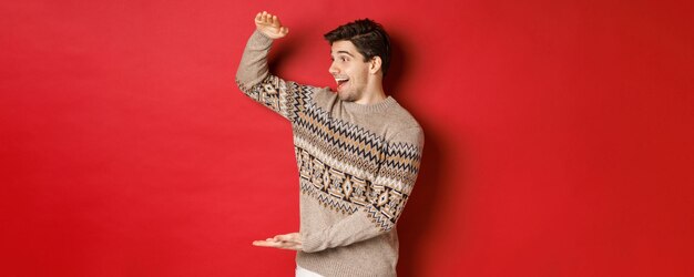 Portrait d'un jeune homme heureux en pull de noël, montrant un grand cadeau, souriant et émerveillé par un cadeau cool, debout sur fond rouge.