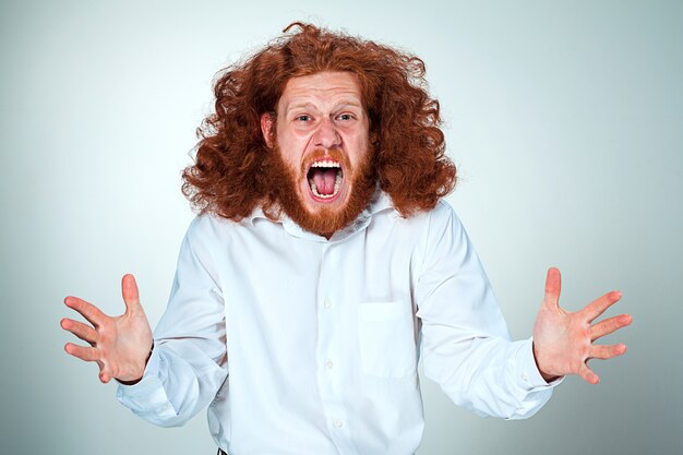 Portrait de jeune homme hurlant aux longs cheveux roux et expression faciale choquée sur mur gris