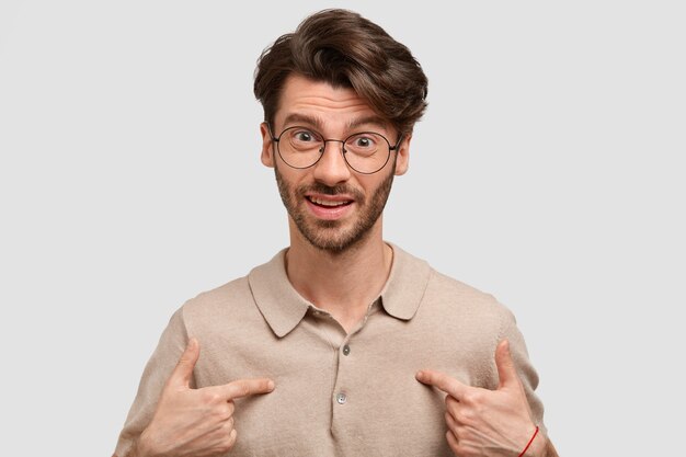 Portrait de jeune homme indigné indique à lui-même, perplexe d'être choisi pour présenter le travail du projet, porte des lunettes, isolé sur un mur de stuido blanc